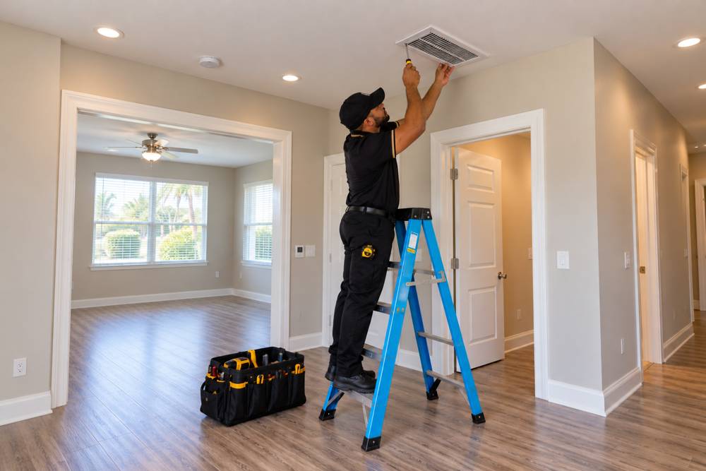 Maintenance professional on a ladder replacing a ceiling vent cover in a bright vacant rental, with a tool bag on the floor